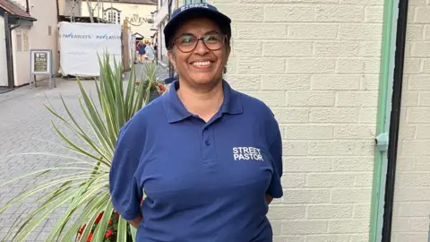 A woman with brown hair is wearing a cap and blue t-shirt that says 'Street Pastor'. She is standing next to a white washed wall.