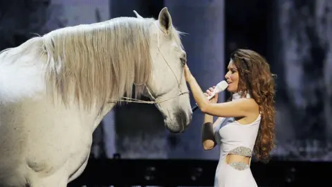 Getty Shania Twain singing, alongside a white horse. 