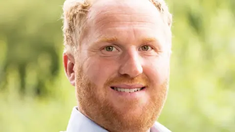 NFU Oli is smiling at the camera. He has a ginger beard and brown eyes. 