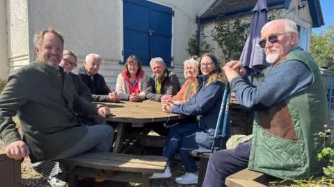Four men and four women sit around an outdoor wooden table in front of a village pub. The pub is rendered with blue shutters at the windows. 