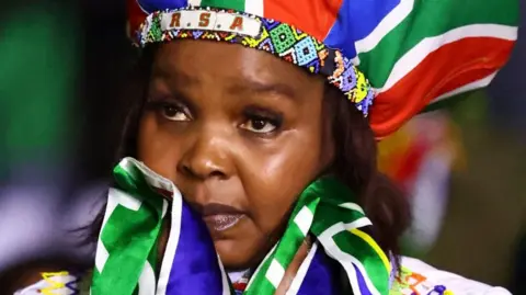 A woman with a hat in South African colours and a beaded headband that says "RSA" looks sad. A South African flag is wrapped round her neck.