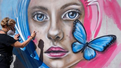 A woman paints a mural of a woman with pink and blue hair