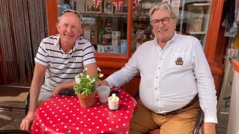 Two men smile while sitting at a small red circular table outside the front of a cafe. A pot plant is on the table along with cups of coffee. The store behind them has an orange frame with historic items in the window.