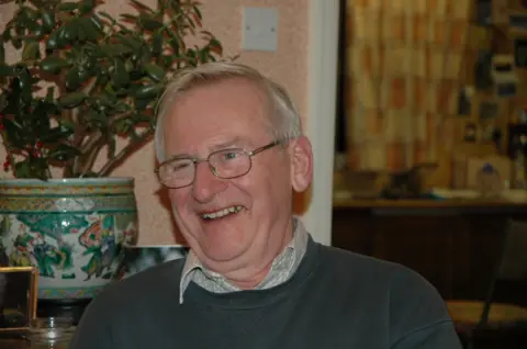 Family handout A smiling Richard Periam is wearing glasses. He has short grey hair and is wearing a checked shirt and green jumper. Behind him is a plant in an Asian-style pot.