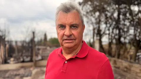 A man with grey hair and a red polo shirt on. He is looking towards the camera with a stern look on his face.