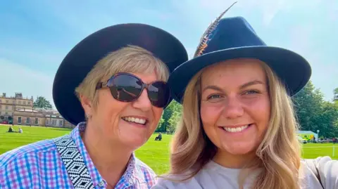 Hayley Vaughan A picture of two blonde women side by side, the woman on the left has short hair with a dark hat on. She is also wearing a blue and pink check shirt and dark sunglasses. The woman on the right has long hair, with a navy blue hat on and is wearing a grey top.