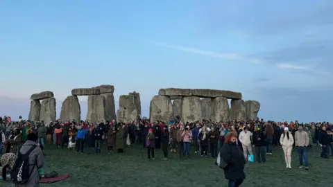 Sophie Parker Stonehenge stones, large stones in a circle, surrounded by large groups of people wrapped up warm in jackets and scarves.