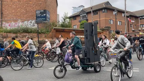 People are riding bikes along a street, some cyclists are flying flags while others have large black speakers on the back of their bikes. There are four-storey homes in the background.