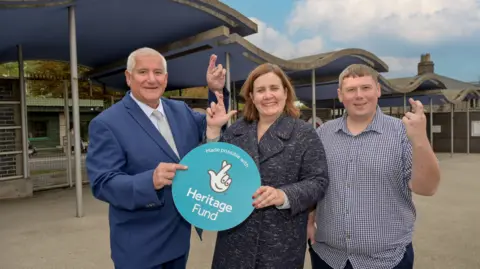 Dudley Council Three people are stood next to each other at the entrance to a zoo. The man on the left has white hair and is wearing a blue suit with white shirt and tie. He has fingers in his left hand crossed and is hold a blue, circular Heritage Fund sign with his right. Also holding the sign, with her left hand, is a woman with long brown hair. She is crossing the fingers on her right hand. She is wearing a patterned long blue coat. To the right stands a man with brown hair, wearing a checked blue and white shirt. He is crossing the fingers of his left hand. They are all smiling at the camera.
