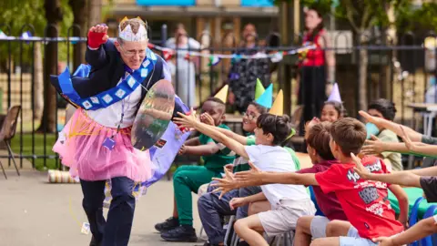 David Lewin is dressed in an assortment of crazy clothing, including a pink tutu and a paper crown. He is running past students whilst doing a Superman-esque pose.