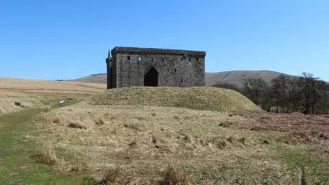 Billy McCrorie The remains of an ruined castle sit on top of a mound of earth in the rolling Borders landscape beneath a blue sky