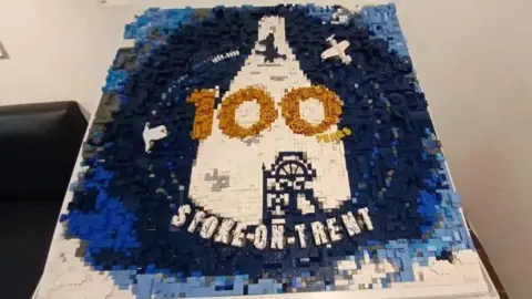 Stoke-on-Trent City Council A Lego model of Stoke-on-Trent's centenary logo, which is made up of hundreds of grey, black, blue and white plastic bricks. Models of a Spitfire plane and a bottle oven can be seen in the middle, alongside the number 100 in gold Lego pieces and the word "Stoke-on-Trent" written in white along the bottom.
