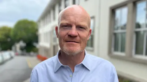 BBC A man looking directly at the camera wearing a blue shirt. Background is blurred but a building on the right can be seen as can a road to its left. Trees and cloudy skies behind.