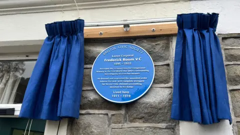 Blue plaque with opened ceremonial curtains on either side. Plaque reads "Bristol Civic Society', Lance Corporal Frederick Room VC, 1895-1932, Awarded the Victoria Cross for conspicuous bravery in the First World War when commanding his company of stretcher-bearers. He dressed and evacuated the wounded under intense fire and with complete disregard for his own life, showed unremitting devotion to his duties. Lived here 1911-1919.