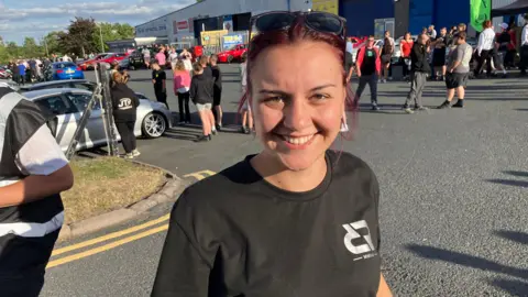 Tia Fenton, wearing a black top with a white small logo on it, smiles into the camera with short red-dyed hair and sunglasses perched on her head. Behind her, groups of people mill around cars.