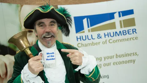 Michael Wood Michael Wood - a man in a green and black town crier uniform - is holding a brass handbell and a mug with "Mouth of the Humber" written on it.