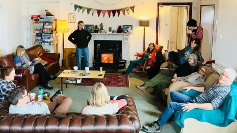 Eleven of the 21 members gather in the communal lounge to chat. There are sofas and chairs of different colours while in the background the fire burns and shelving filled with games can be seen. 