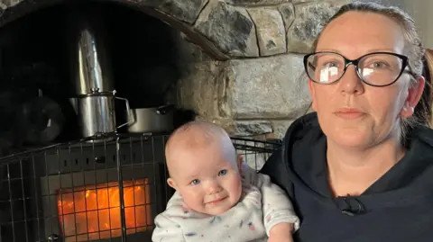 Nicola Graham, who is wearing a black hoodie and black-rimmed glasses and has a brown ponytail, sits next to a black woodburning stove with a chrome chimney. She is holding her daughter Neavh, an 8-month-old baby wearing a white babygrow.