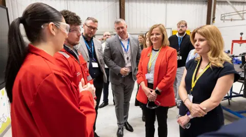 North East mayor Kim McGuinness and North Tyneside mayor Karen Clark visiting the Newcastle College Energy Academy in Wallsend. They are standing in a large room and talking to two students dressed in red overalls and wearing safety goggles, while other people watch them.
