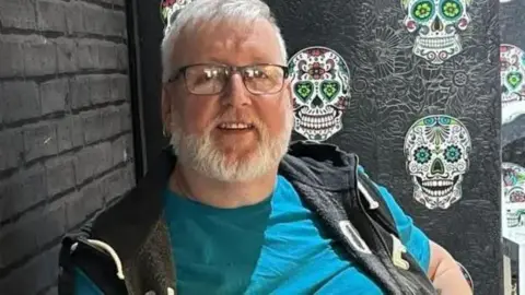 Family handout Kevin Price, who had white hair and a beard, is photographed wearing dark glasses. He smiles as he is photographed sitting in a room with black wallpaper, wearing a gilet on top of a blue t-shirt.
