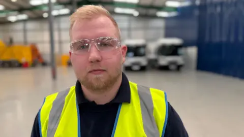 BBC A man with strawberry blonde hair and blonde facial hair is wearing clear protective glasses and is wearing a black t-shirt with a 