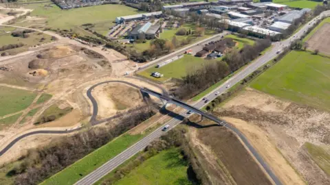 Urban&Civic An aerial shot of a footbridge spannning the A10 near the site of the new town at Waterbeach in Cambridgeshire, with the road cutting through the landscape from bottom left to top right. On either side of the single carriageway road are fields and trees or hedgerows.