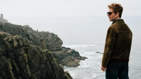 Actor Damien Molony in character as Jim Bergerac. He looks over part of Jersey's coastline. He has a brown coat and dark sunglasses. Rugged rocks are dotted along the coast. It is a gloomy day as waves crash into the rocks.