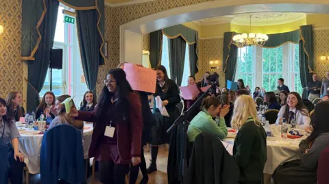 A room with several round tables and young people sat around them. In the middle, seven teenage girls are walking through the room, holding pink and green posters, one of which says 'Make America Green Again'.