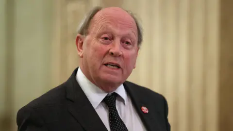 TUV leader Jim Allister is an older man with fair hair. He is wearing a dark jacket and tie which is black with white spots and a white shirt. There is a badge on his left lapel which is red with white lettering. He is standing in front of a beige background.