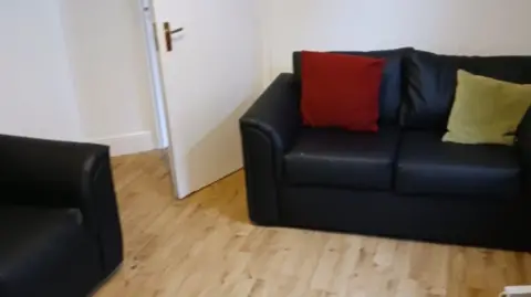 Restore/Leslie Midwinter A tidy living room with two black sofas. One of the sofas has two cushions, one red, one yellow. The room is very tidy and has minimal items. 