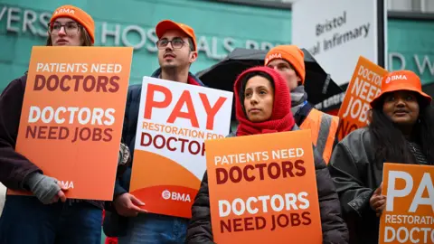 People holding placards saying 'patients need doctors, doctors need jobs' stand outside wearing hats and thick clothes outside Bristol Royal Infirmary on Wednesday.