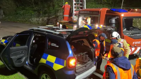 BBC A Guernsey coastguard car, with its back left door and boot open. There are people in coats and orange hi-vis vests. There is a red Guernsey Fire and Rescue truck. Both are parked facing different directions. The road is grey. There is green grass to the left of the Guernsey Coastguard car. 