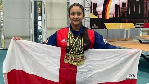 Kaifah Imran pictured holding up a large red and white St George's England flag. She has long black hair and six gold medals around her neck and wears a blue and red jacket. She is standing in the middle of a gym.
