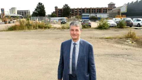 LDRS Philip wears a blue checkered suit and blue tie and is standing on the brownfield site which backs on to a car park. There are buildings in the background and metal barriers with bits of rubbish blown into them.