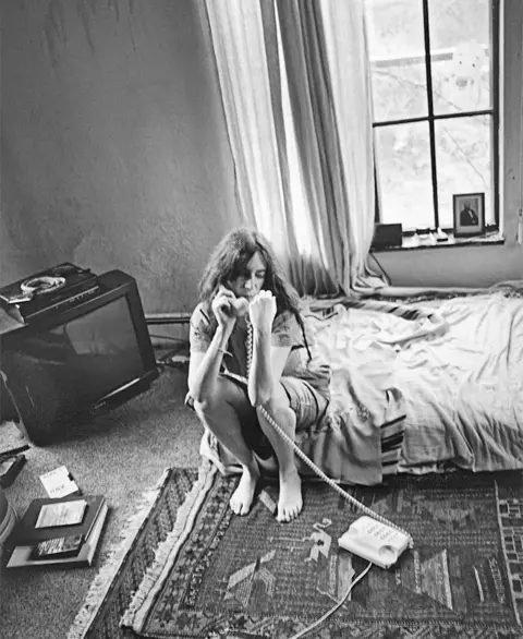 Steven Sebring Patti Smith in T-shirt and shorts, sitting on the edge of an unmade bed with receiver in right hand and phone on a large rug.
