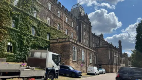 Picture of the Derbyshire County Council building, which is built of stone with several vans and cars parked outside. 