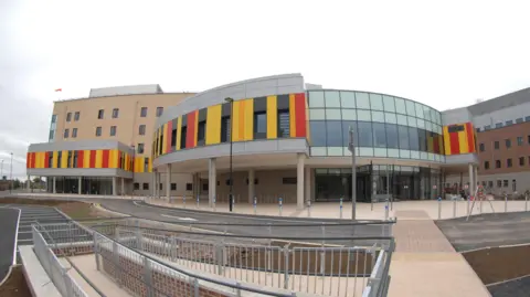 A photograph taken outside Royal Stoke Hospital showing yellow, black and red vertical panels alongside windows.