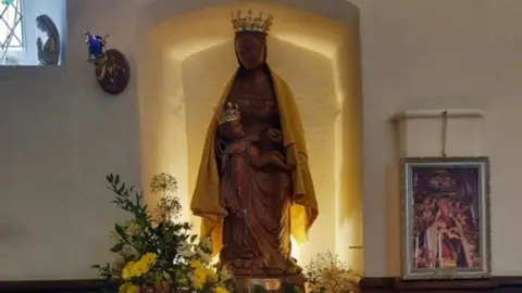 Phil Roberts A replica of a statue of Our Lady of Grace from Nettuno standing in a back-lit niche inside St Mary at the Elms in Ipswich. It is crowned, wearing a cloak and holds a crowned infant Christ on its lap. The is a flower arrangement on the left and candles in front of it.