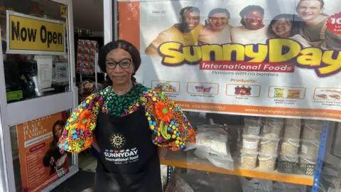 A woman in a black apron which says "sunny day international food". She is wearing a bright and multi-coloured top and is stood in front of her store with the sign "sunny day international foods" outside.
