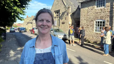 Charlotte Fereday smiles for the camera as she stands outside the stone Victorian school building. She has tied back hair and is wearing denim dungarees and a blue shirt.