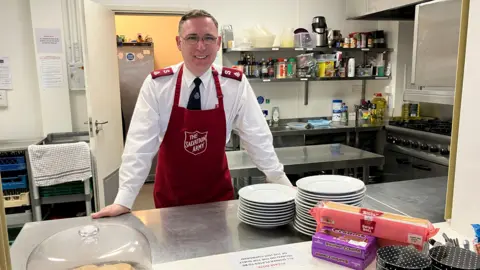 Cianna McNally A man wearing a white shirt and black tie stood behind a food counter. He is also wearing a dark red apron with the words the Salvation Army written on the front. He is in a kitchen area and stood next to plates and food such as cake and biscuits with kitchen utensils and an oven behind him.