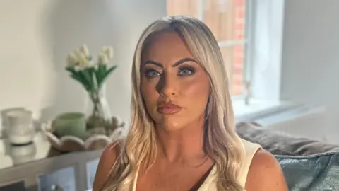 A woman with long blonde hair and blue eyes is looking directly into the camera, unsmiling. She is wearing a light-coloured sleeveless v-neck top. The background is out of focus but she is sitting on a grey solfa with light green cushions, behind her is a table with white flowers on it and a window. 