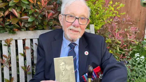 BBC Alan Kennett holding up a picture of himself during 1942 in the RAF. He is wearing glasses and has a white beard.