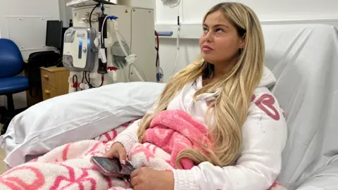 Kenzi Quinn sits in the Renal Unit at Sunderland Royal. She is sitting up in a bed and connected to a dialysis machine. Kenzi has blonde hair and is wearing a white hooded jumper while wrapped in a pink and white blanket, while holding her iPhone in her right hand. 
