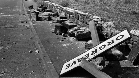 PA Media A black and white photo from 1984 of the Orgreave town road sign on the floor in a pile of rubble. A wall next to a road is torn down and the sign in lying on stones from the broken wall.