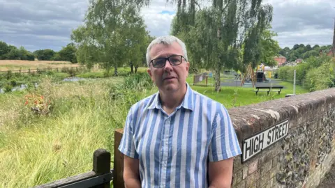 Tony Fisher/BBC Robert Gill, in a stripy short-sleeved shirt, looking concerned as he stands close to the River Misbourne