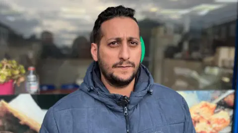 Sophia Seth/BBC A man with dark hair and a beard. He is wearing a dark blue coat and is standing in front of a shop window.