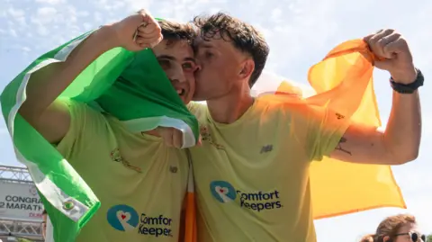 The brothers embrace one another at the end of their Ireland running challenge. Both of them have an Irish flag draped over their shoulders and Jordan kisses Cian on the cheek.