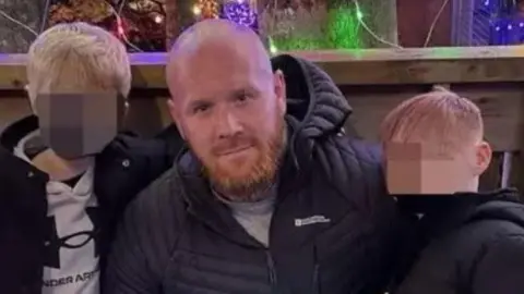 Lenny Scott, who is bald and has a ginger beard, poses with his arms around his young sons. Their faces have been blurred.