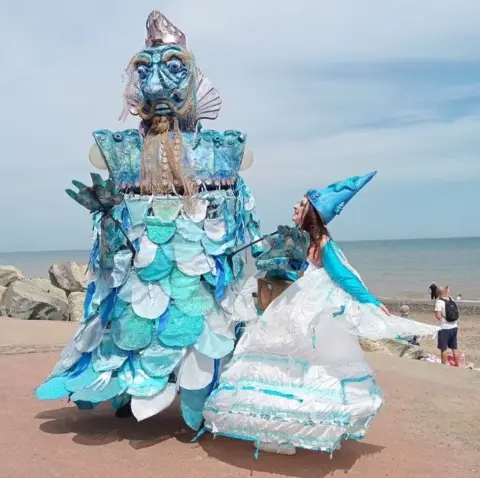 Apus Productions A large puppet a monstrous blue face and shimmering blue, sea-like costume, stands next to a woman wearing a matching outfit while standing on a sea front.